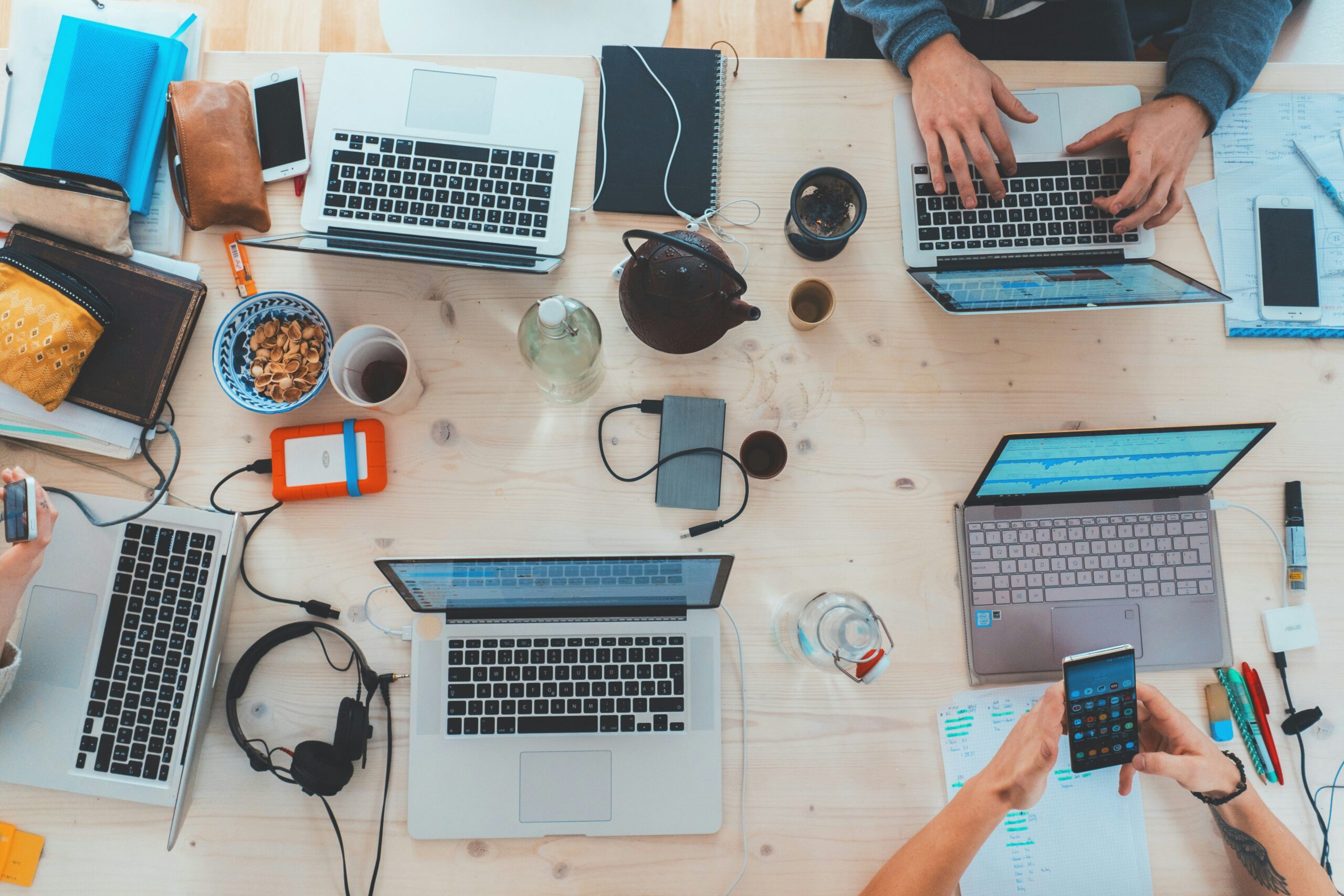 A top-down view of a light wooden table cluttered with multiple laptops, smartphones, notebooks, coffee cups, and electronic accessories being used by people.