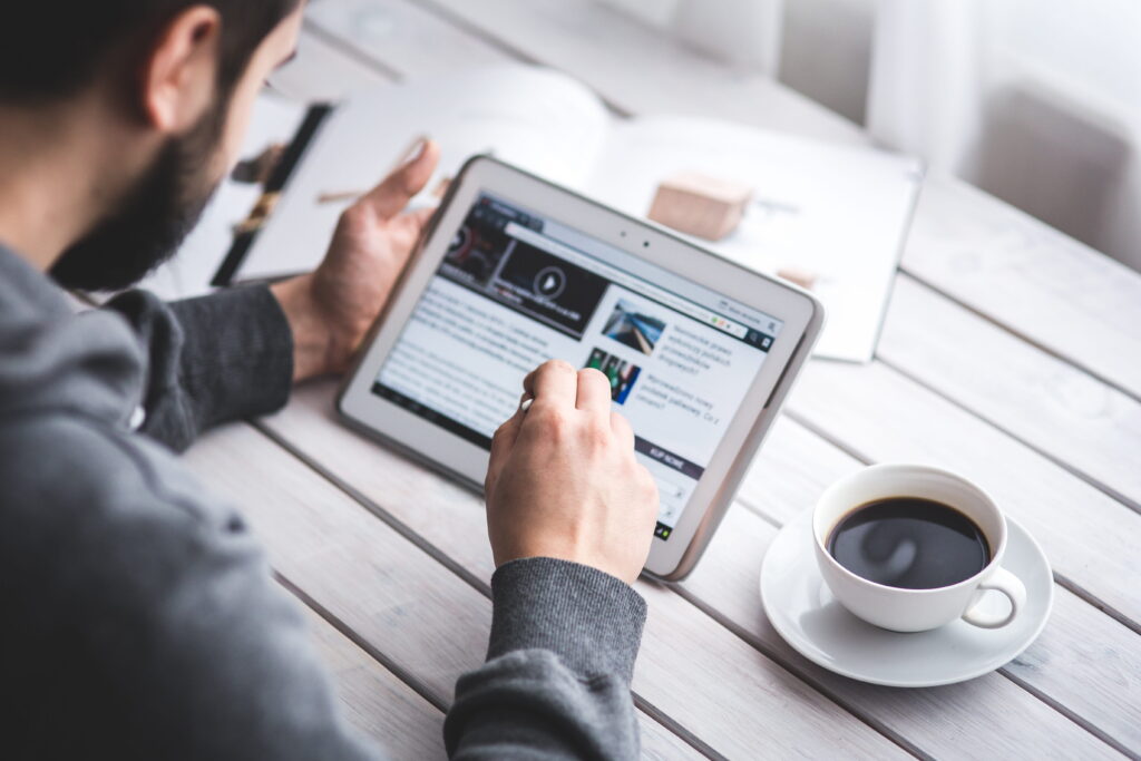 Over-the-shoulder view of a person using a stylus to browse video and news content on a white tablet at a wooden desk next to a cup of coffee.