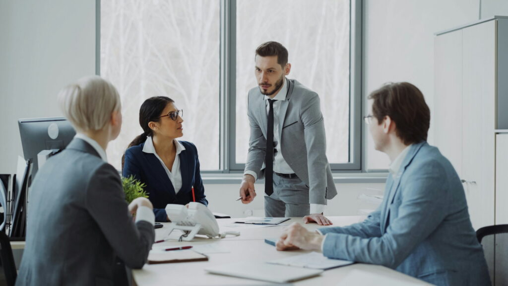 A group of business professionals in formal attire having a meeting around a conference table in an office, with a man standing and leaning over the table while speaking to his colleagues.