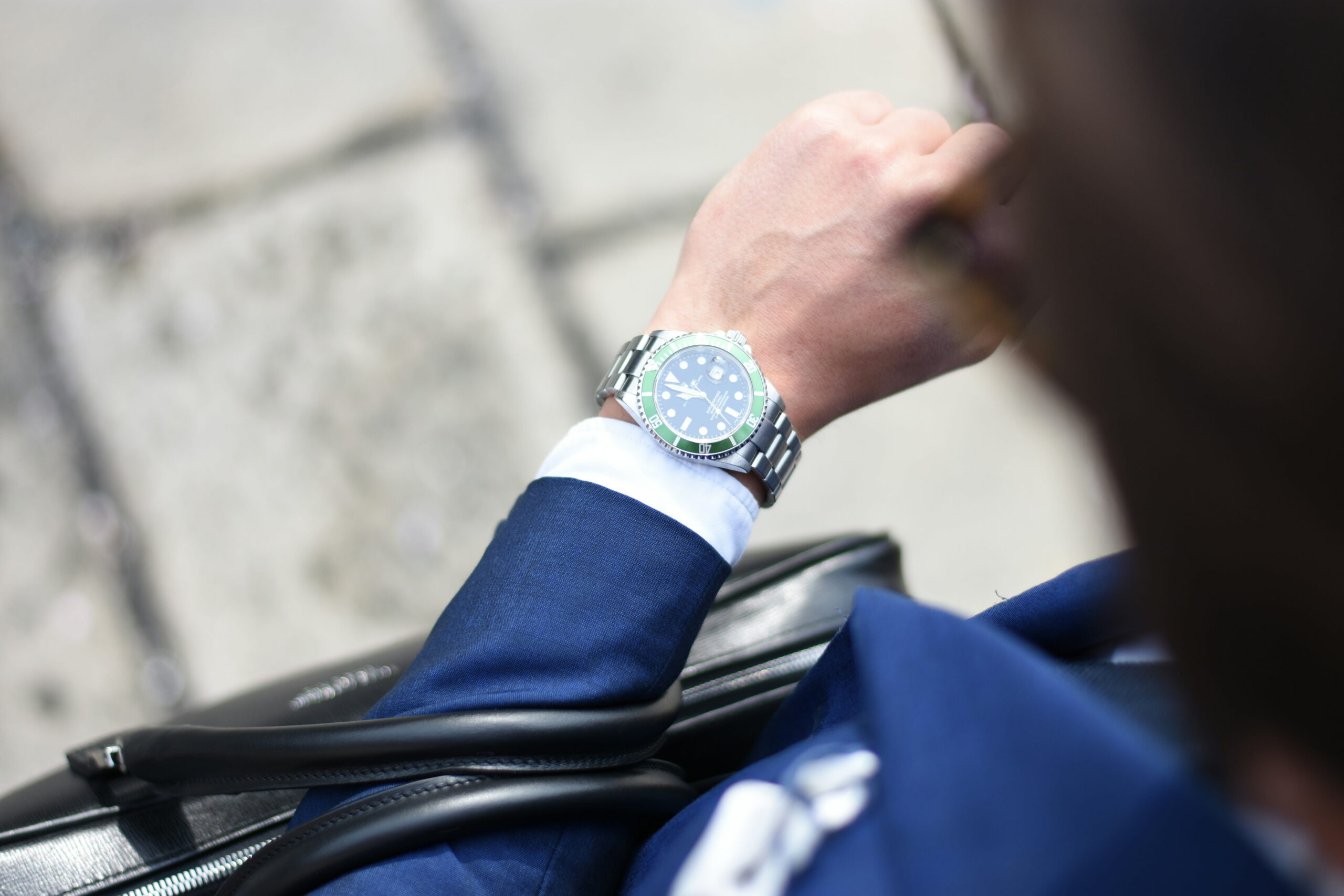 A close-up view of a professional in a blue suit looking at a luxury green-bezel watch on their wrist while holding a briefcase.