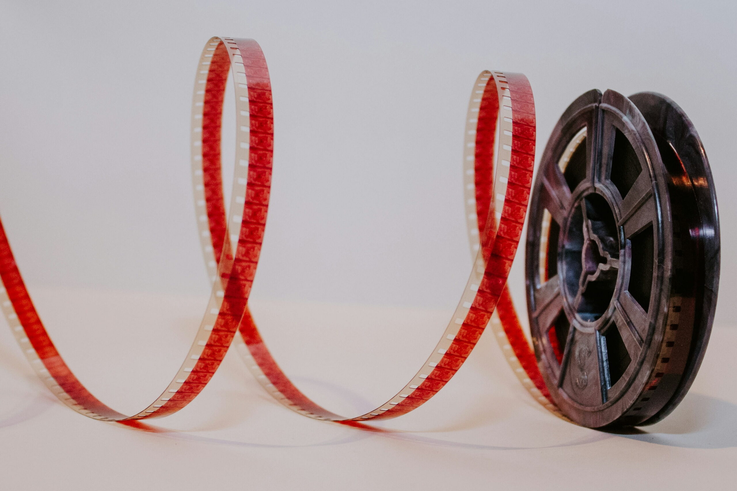 A dark vintage film reel resting next to a long, curved strip of red-tinted motion picture film on a light surface.