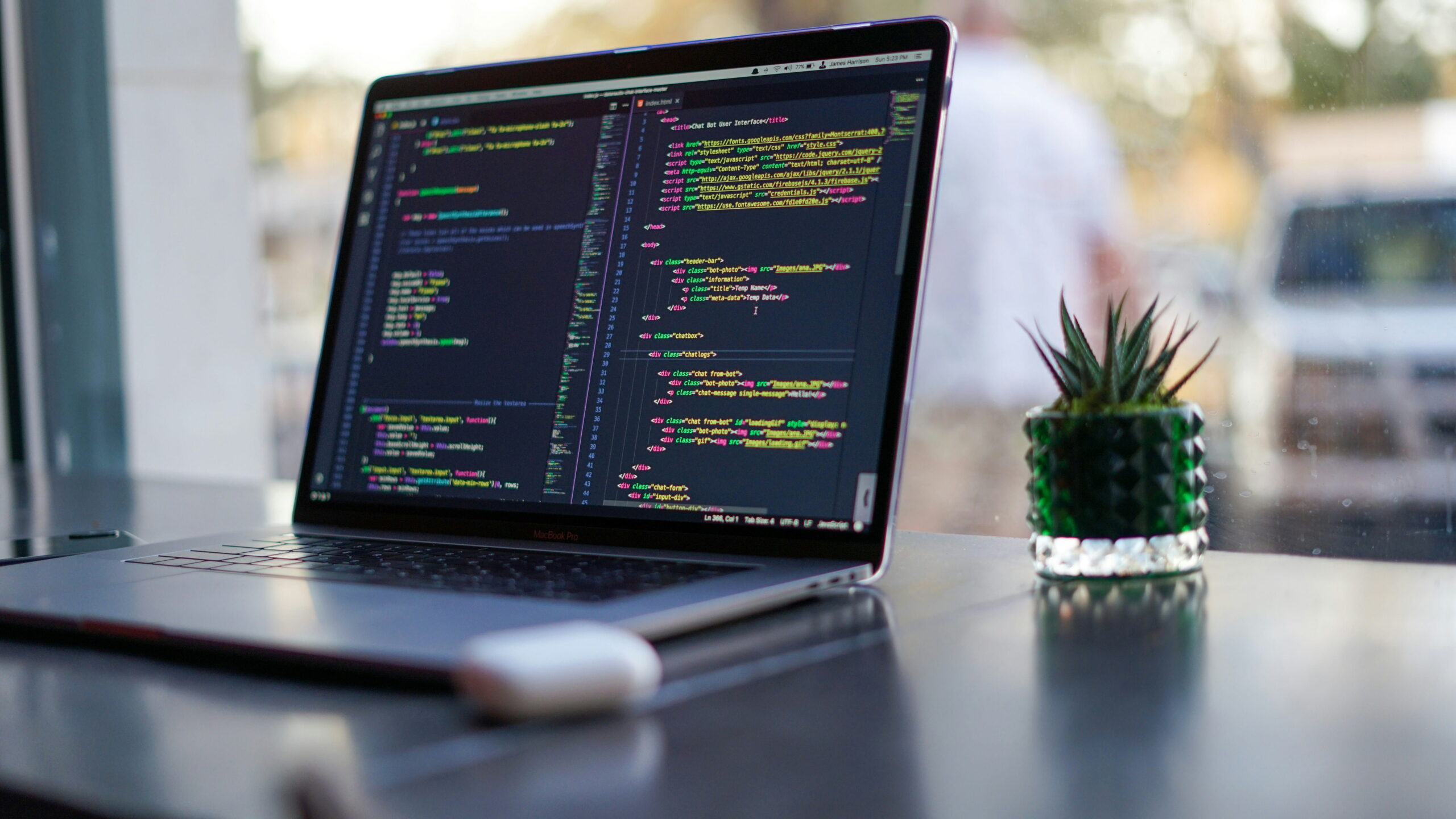 An open laptop displaying lines of programming code on its screen, set on a dark desk next to a small potted succulent and a pair of wireless earbuds.