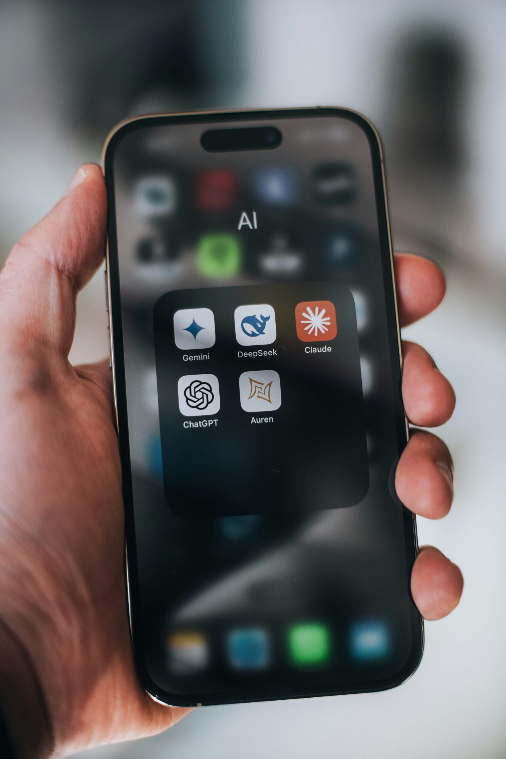 A close-up of a person holding a smartphone displaying an "AI" folder on the home screen, featuring app icons for Gemini, DeepSeek, Claude, ChatGPT, and Auren.
