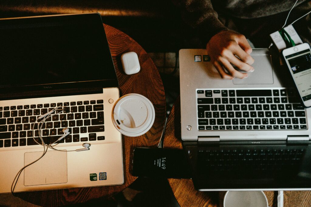 An overhead, close-up shot of two laptops, a coffee cup, wireless earbuds, and a smartphone on a dark wooden table.