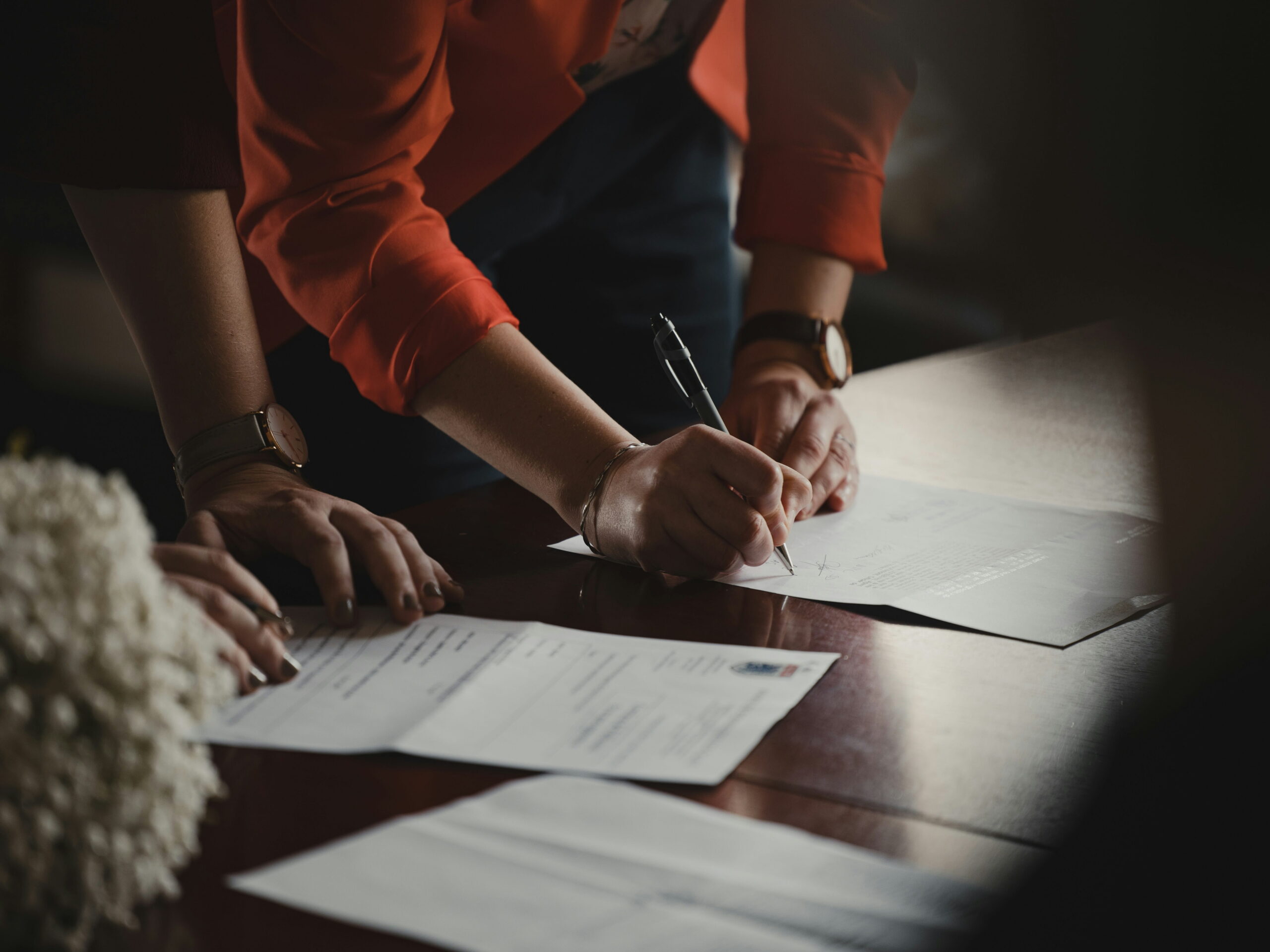 Two people in professional attire leaning over a dark wooden desk to review and sign contract documents with a pen.