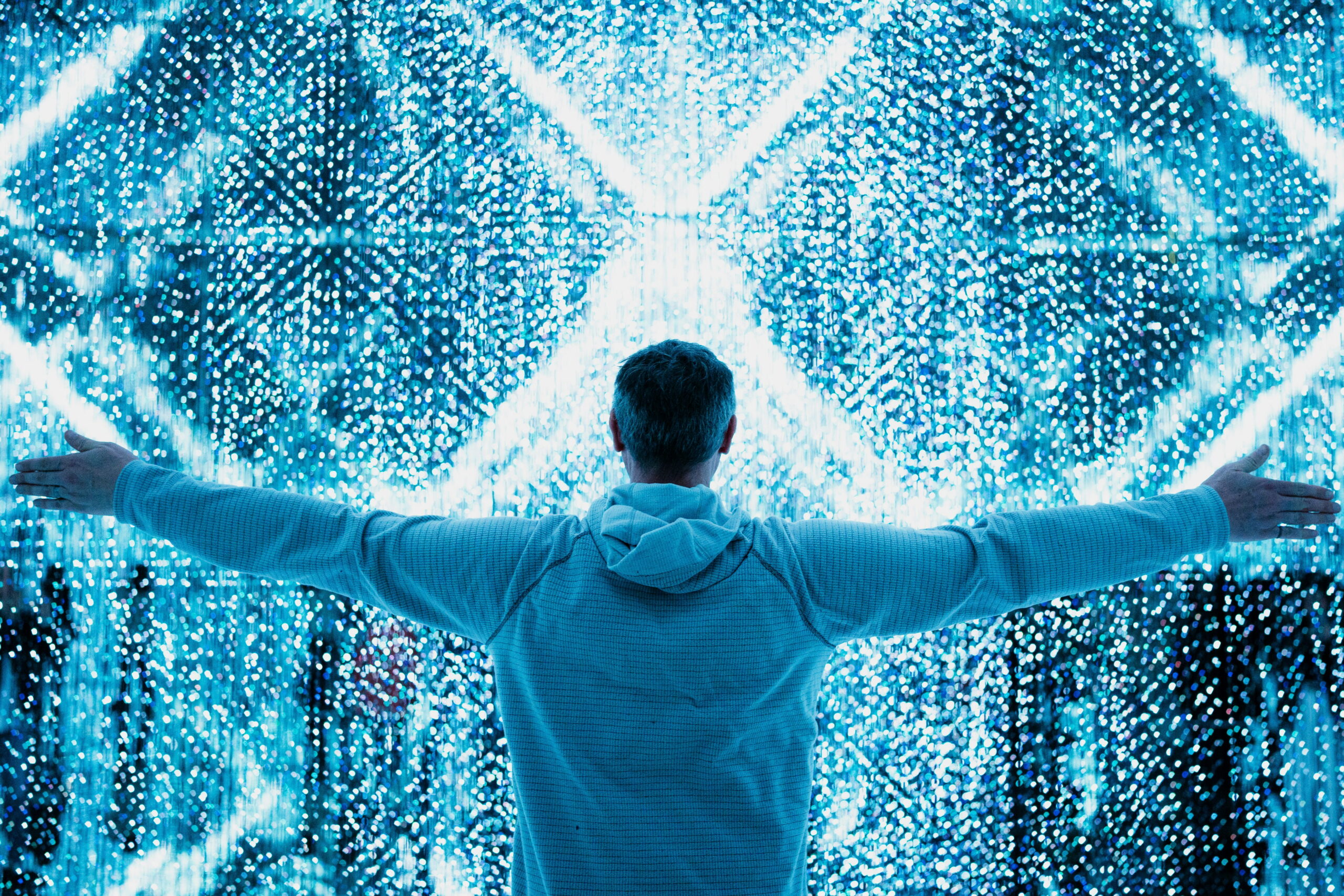 A person from behind standing with their arms outstretched in front of a massive, shimmering wall of bright blue lights and geometric digital patterns.