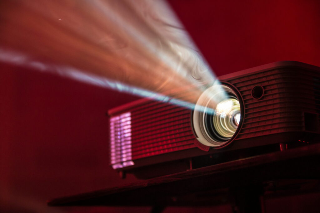 A digital projector projecting a bright beam of light filled with dust particles against a dark red background.