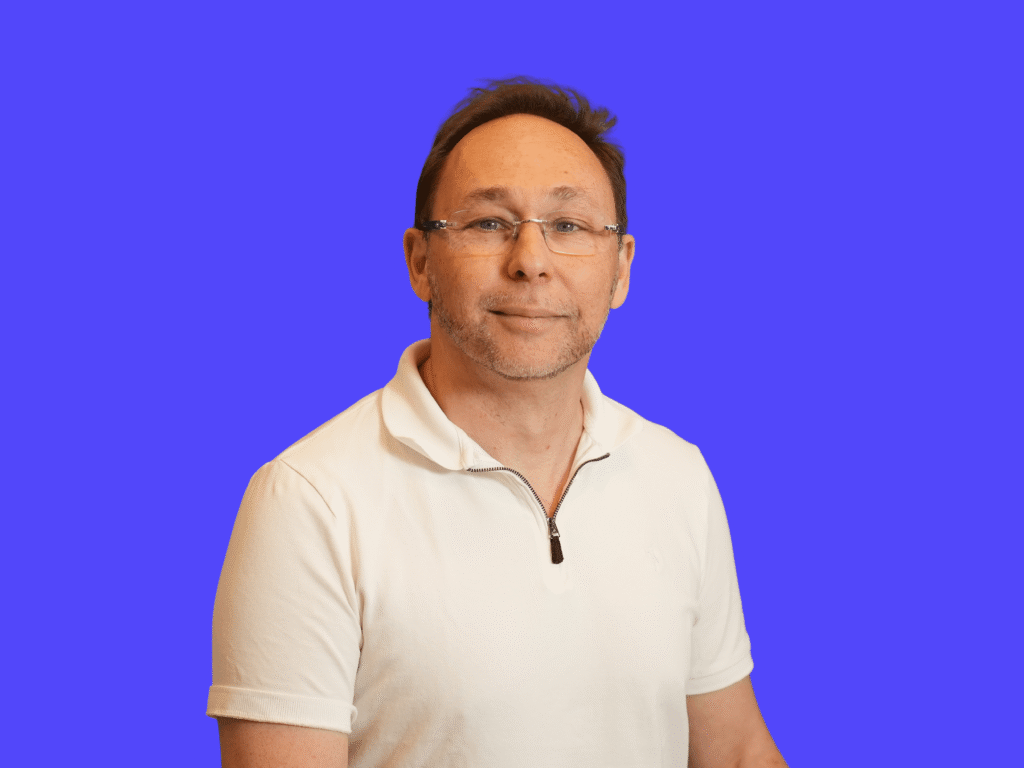 This image shows a man with short brown hair and glasses, wearing a white zippered polo shirt against a solid blue background.