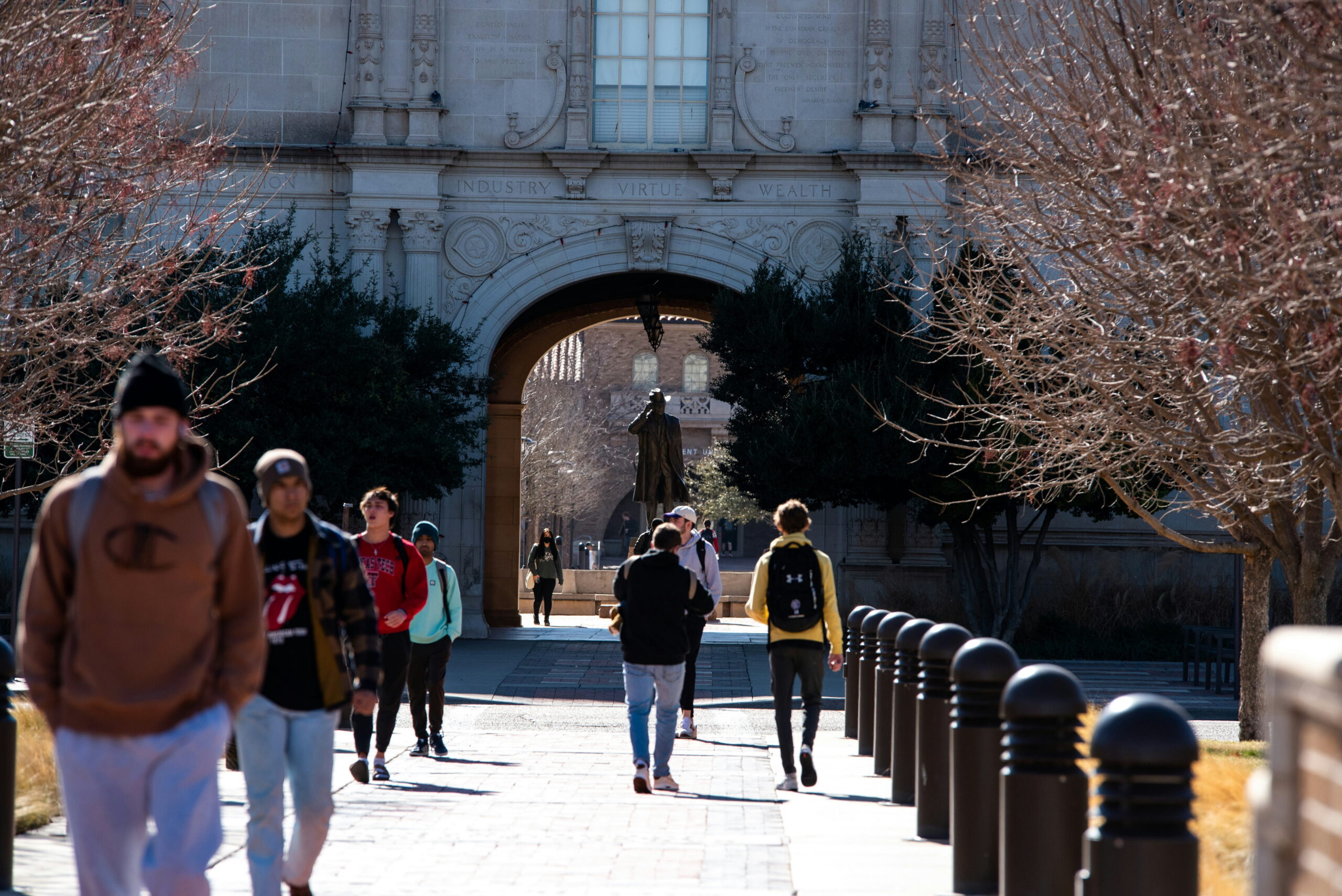 Students walk along a brick pathway on a college campus toward a large stone archway under bare trees.