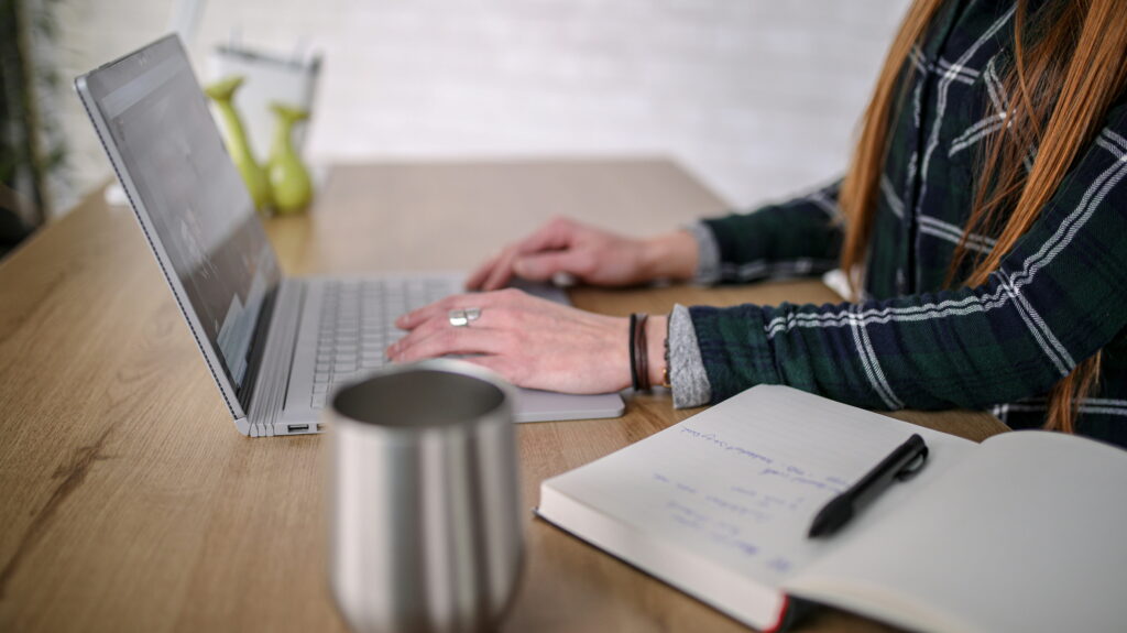 A person with long red hair wearing a plaid shirt types on a laptop at a light wood desk, with an open notebook, a pen, and a metallic mug nearby.