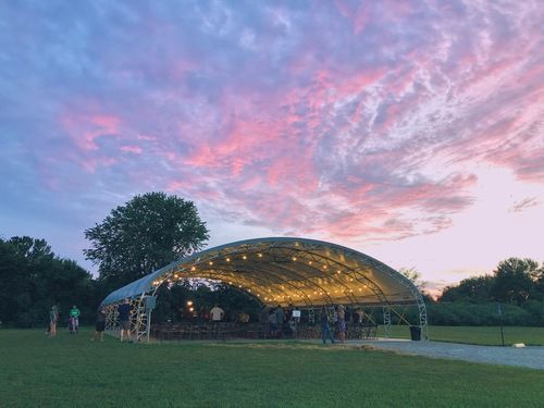 An outdoor gathering space featuring a large, arched metal-framed pavilion lit with string lights, set against a vibrant pink and purple sunset sky over a grassy field with trees.
