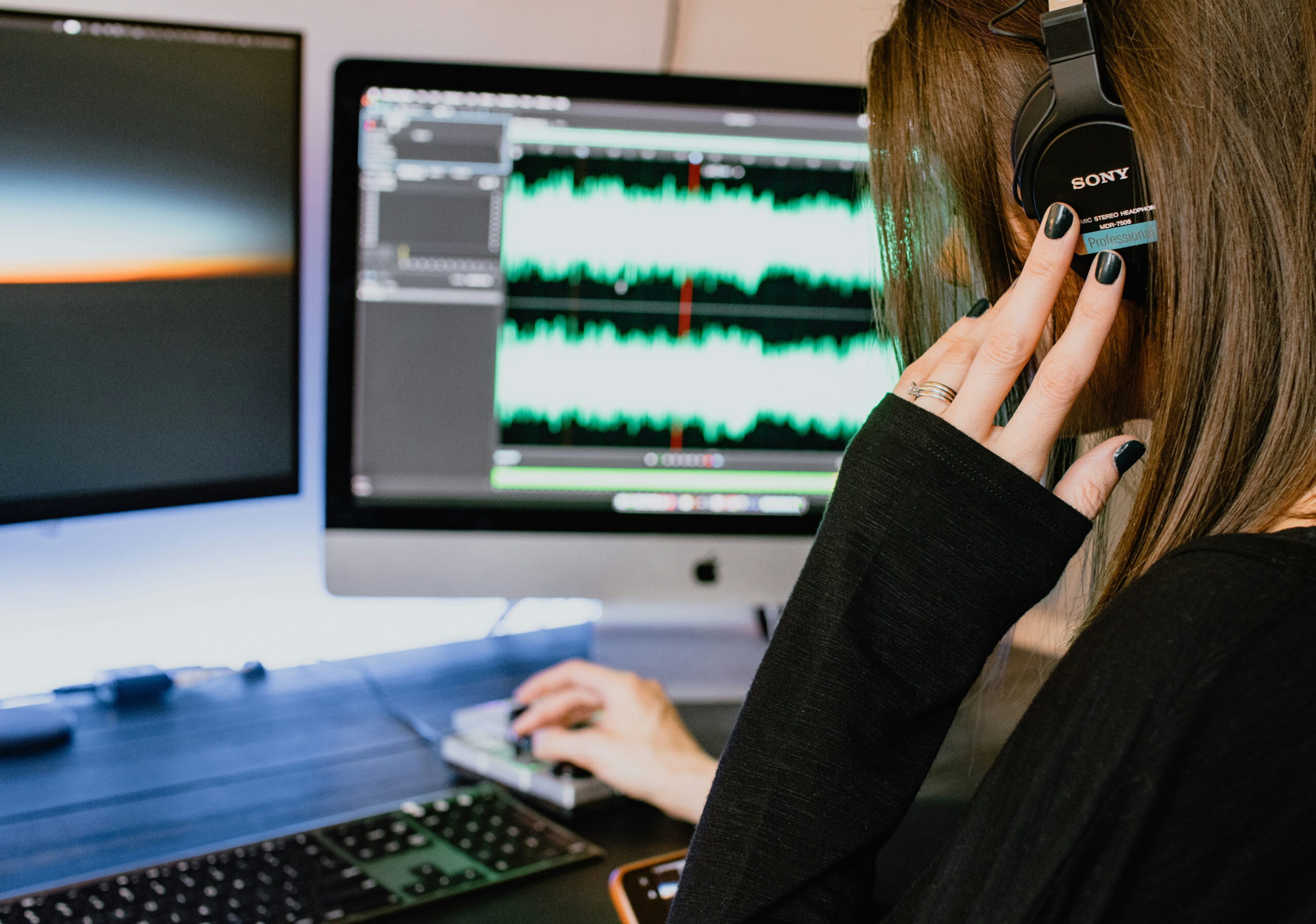 A woman wearing Sony professional headphones adjusting her earpiece while looking at dual computer monitors displaying bright green audio waveforms.