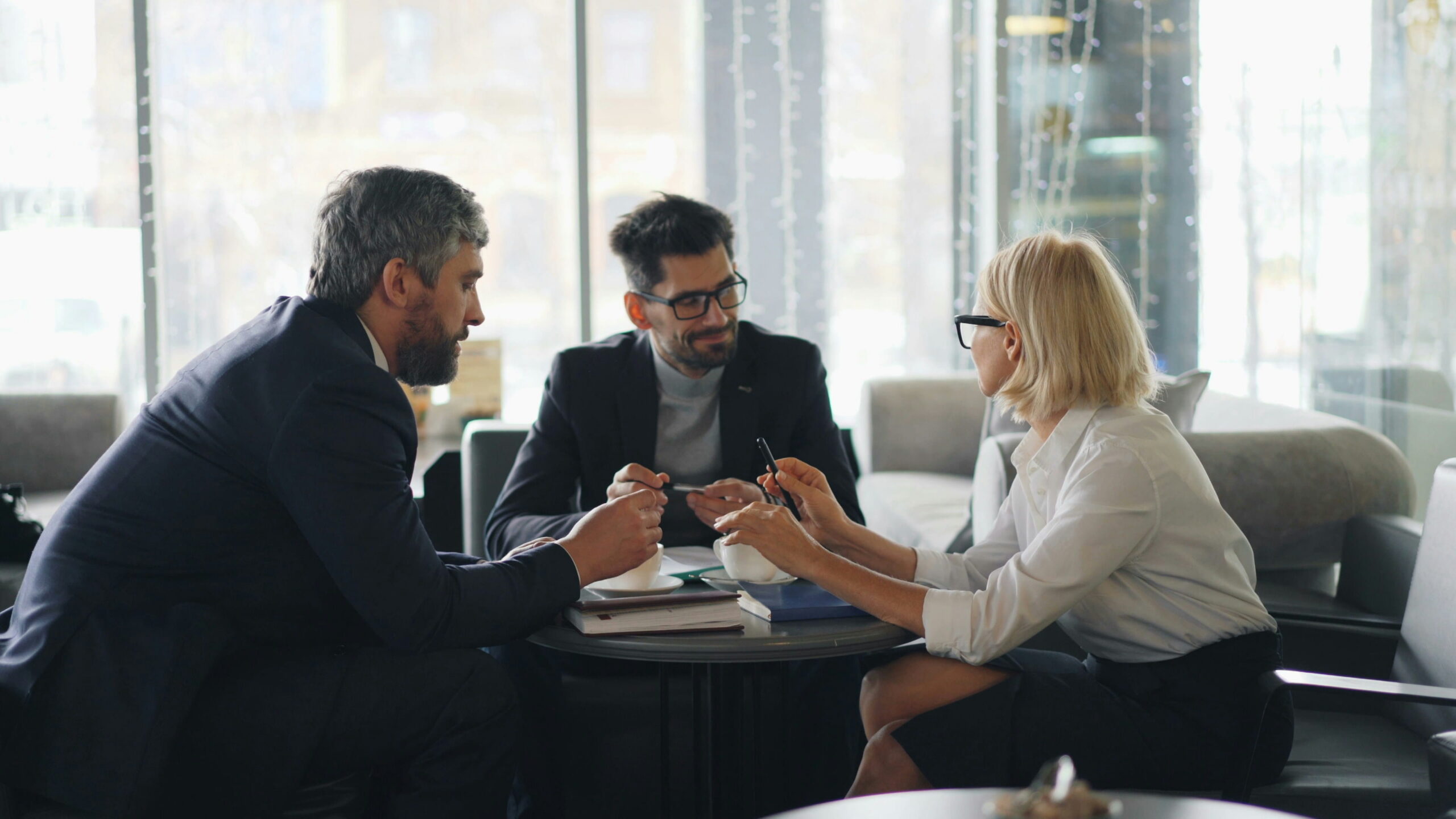 Three professionals in business attire holding a discussion seated around a small coffee table in a contemporary office lounge area with large windows.