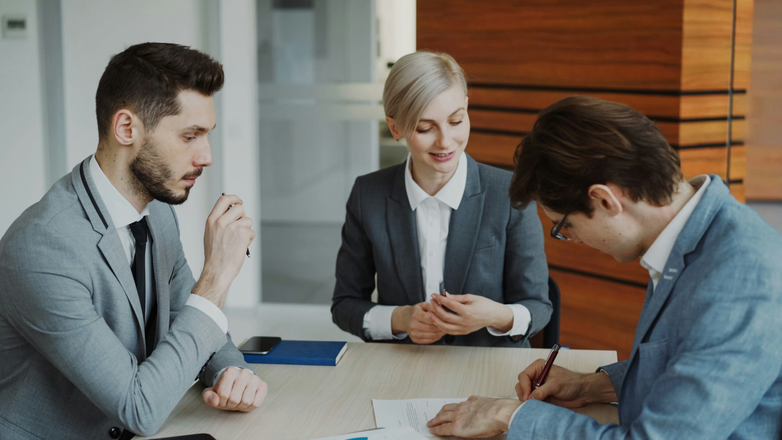 Three business professionals in a modern office, with a man signing a document at a table while a woman and another man look on advising him.