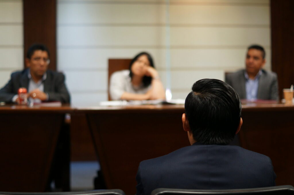 A back view of a person in a suit sitting in a deposition hearing, facing a panel of attorneys and officials seated across a long wooden table.