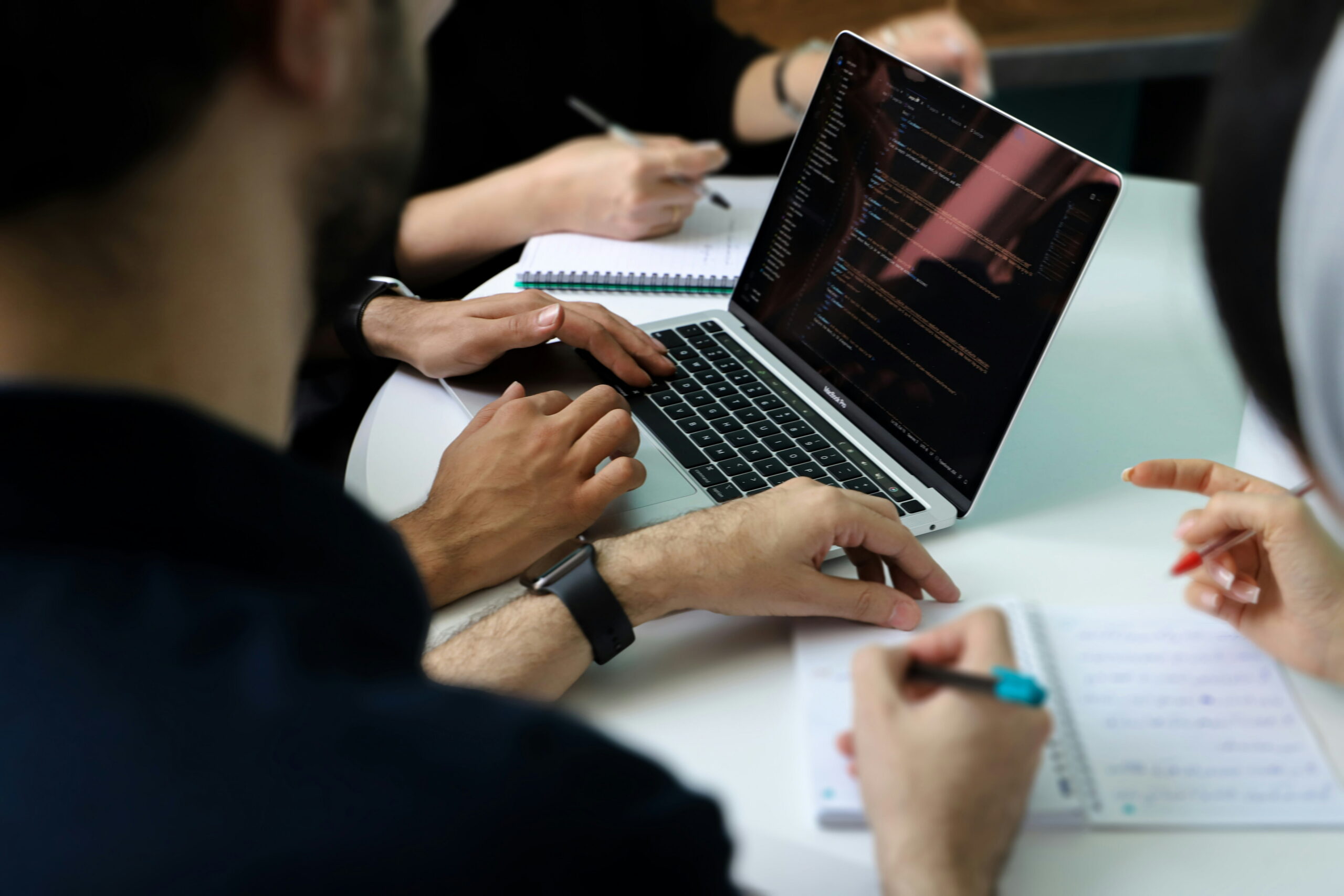 Close-up view of a software developer's hands typing on a laptop computer displaying code lines while team members take notes on notebooks during a meeting.