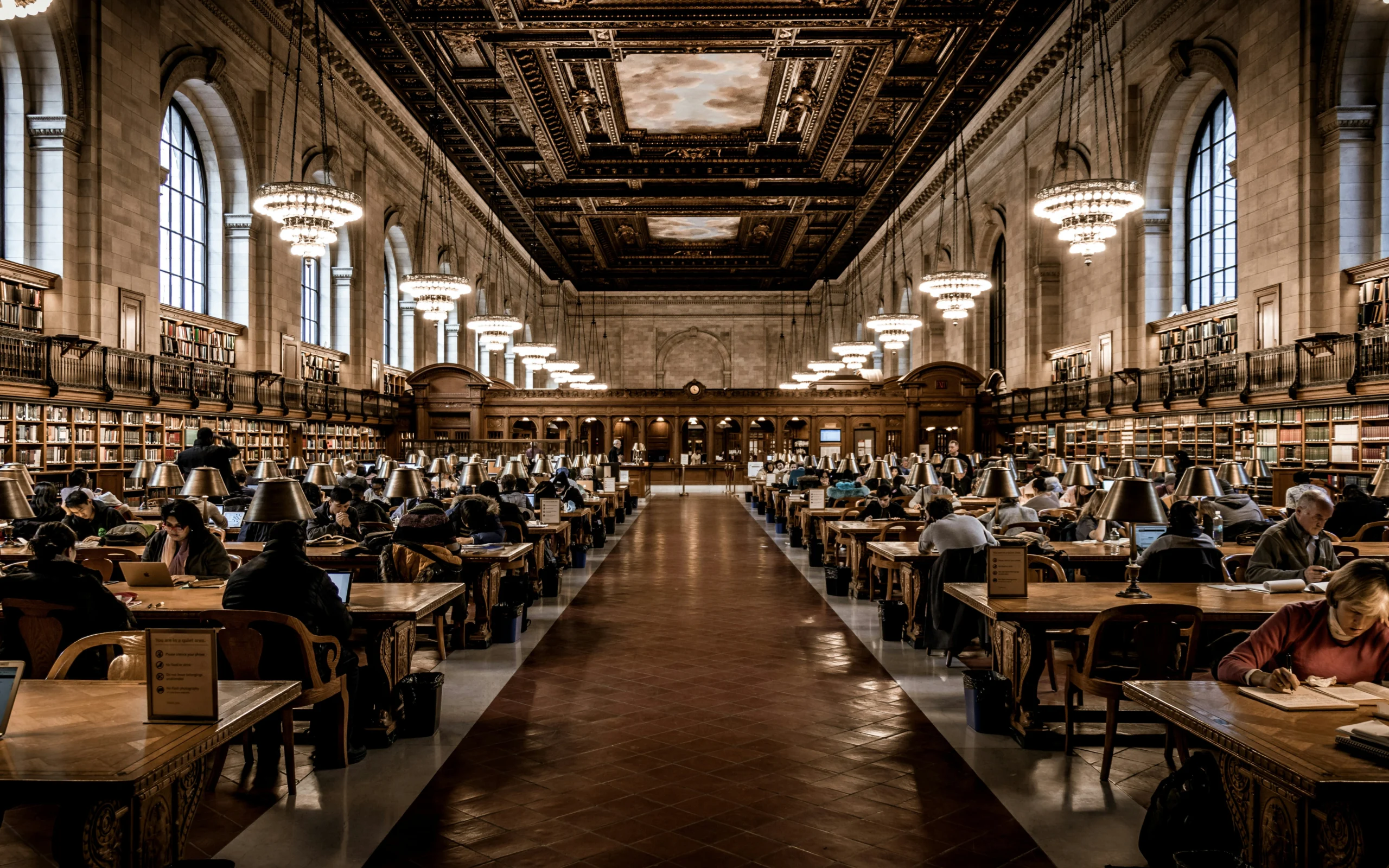 A wide-angle view of a grand, ornate library reading room with rows of wooden study tables lit by green lamps, tall arched windows, bookshelves lining the walls, and an elaborate dark wood and gold ceiling with multiple chandeliers.