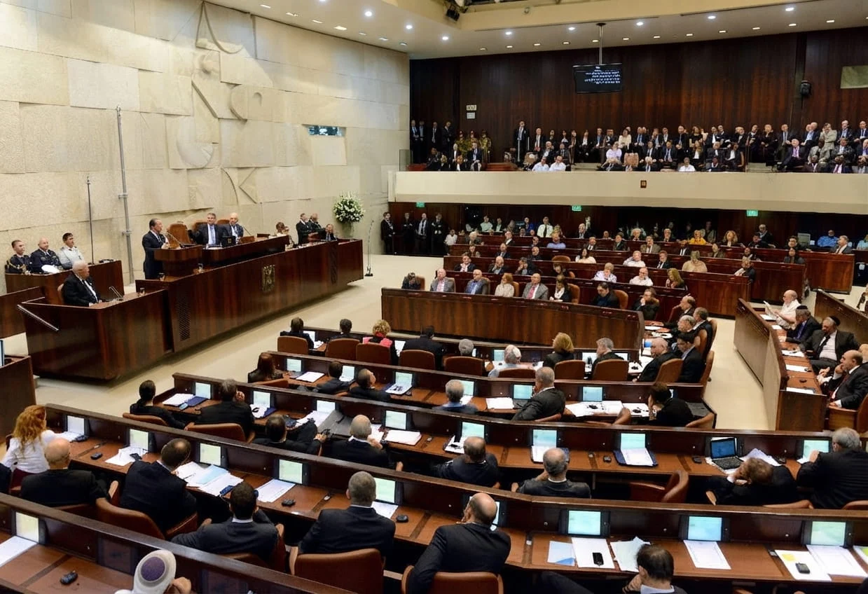 Wide-angle view of the Israeli Knesset parliament chamber filled with seated members, wood-paneled desks, and tiered seating, with the speaker's podium visible on the left.