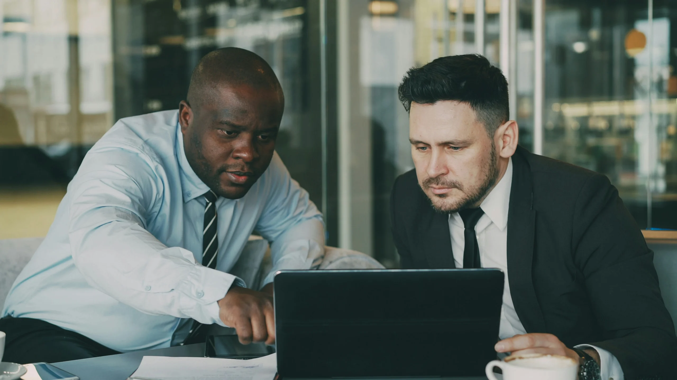 Two business professionals in suits reviewing data and software on a laptop together in a modern office environment.