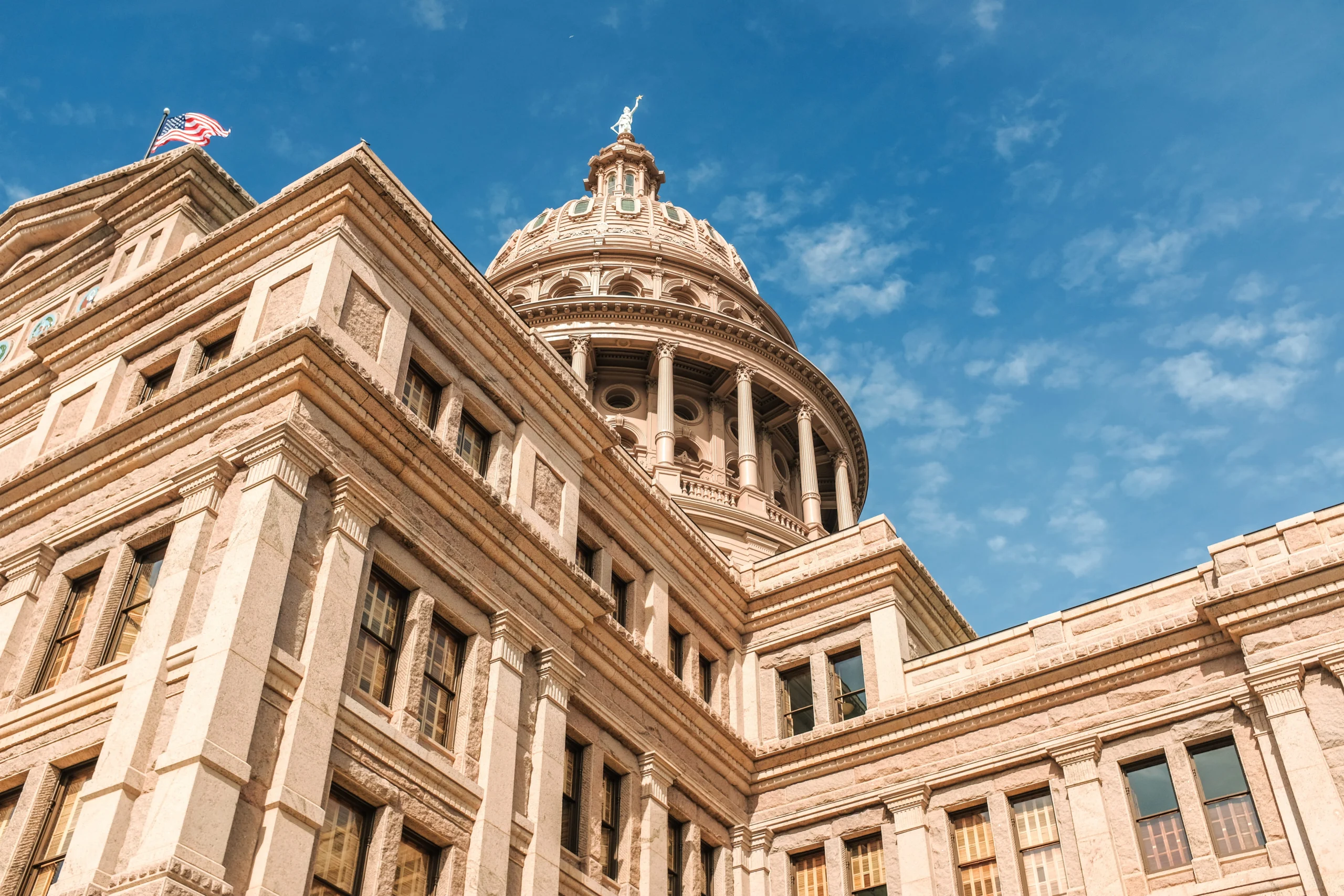 Exterior low-angle shot of the granite Texas State Capitol building in Austin, featuring its prominent dome and architectural details against a bright blue sky with scattered clouds.