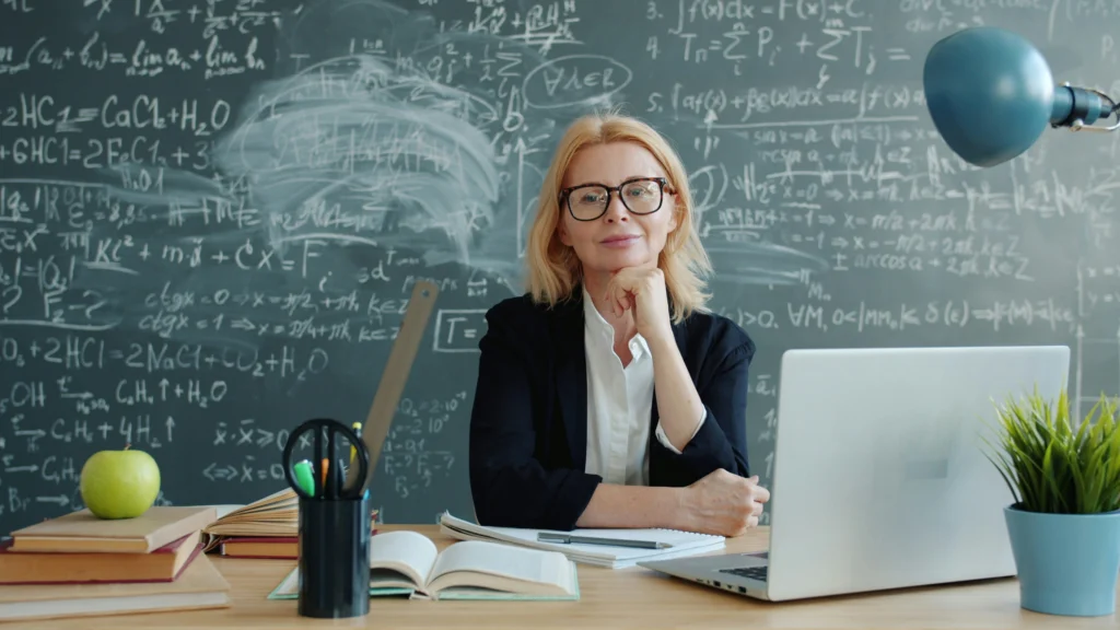 A female teacher in glasses sits at her desk with a laptop and books, looking calmly at the camera in front of a chalkboard of equations.