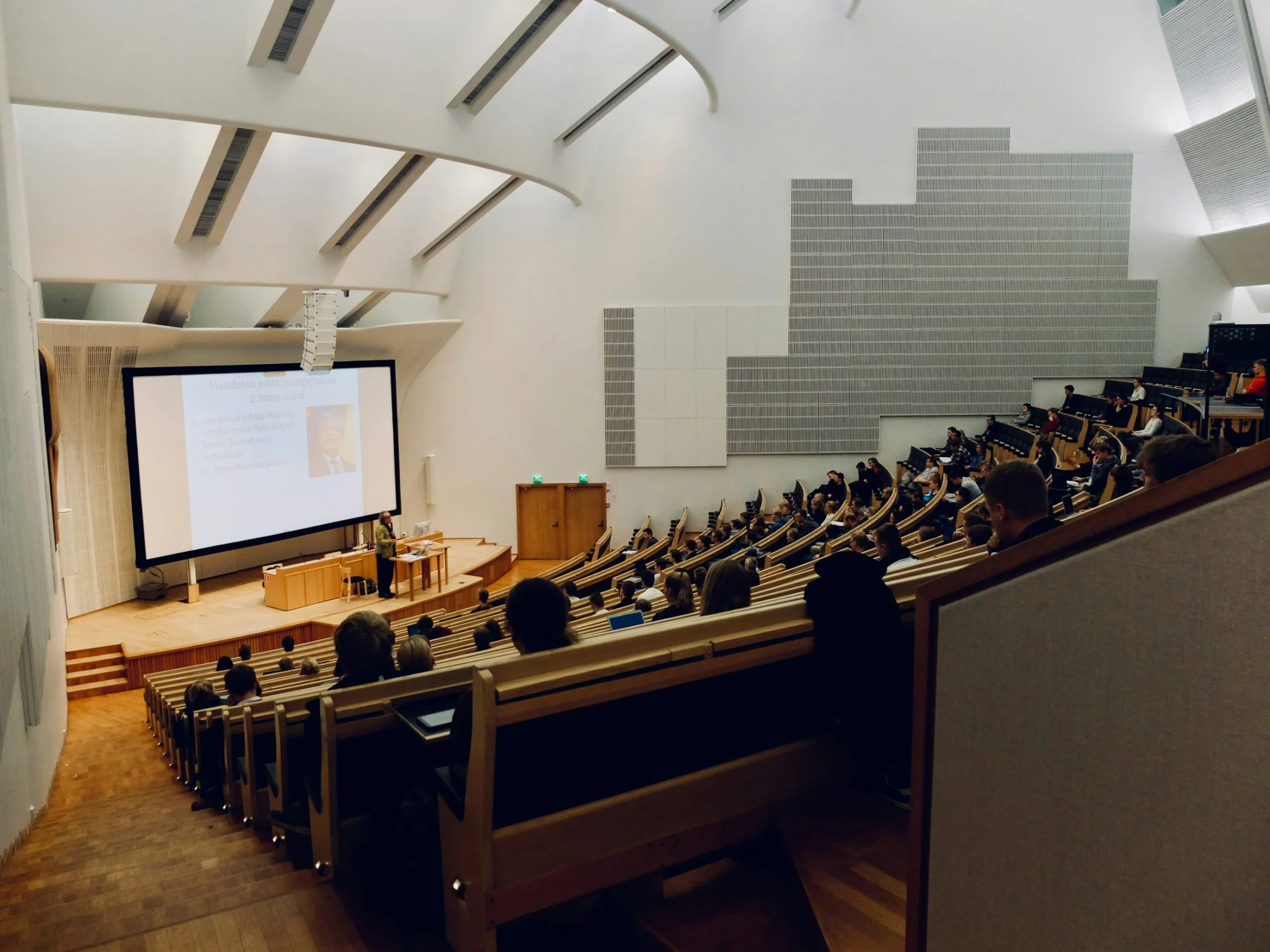 An elevated view looking down into a modern, tiered university lecture hall filled with students seated at wooden desks, facing a large projection screen at the front where a speaker stands.