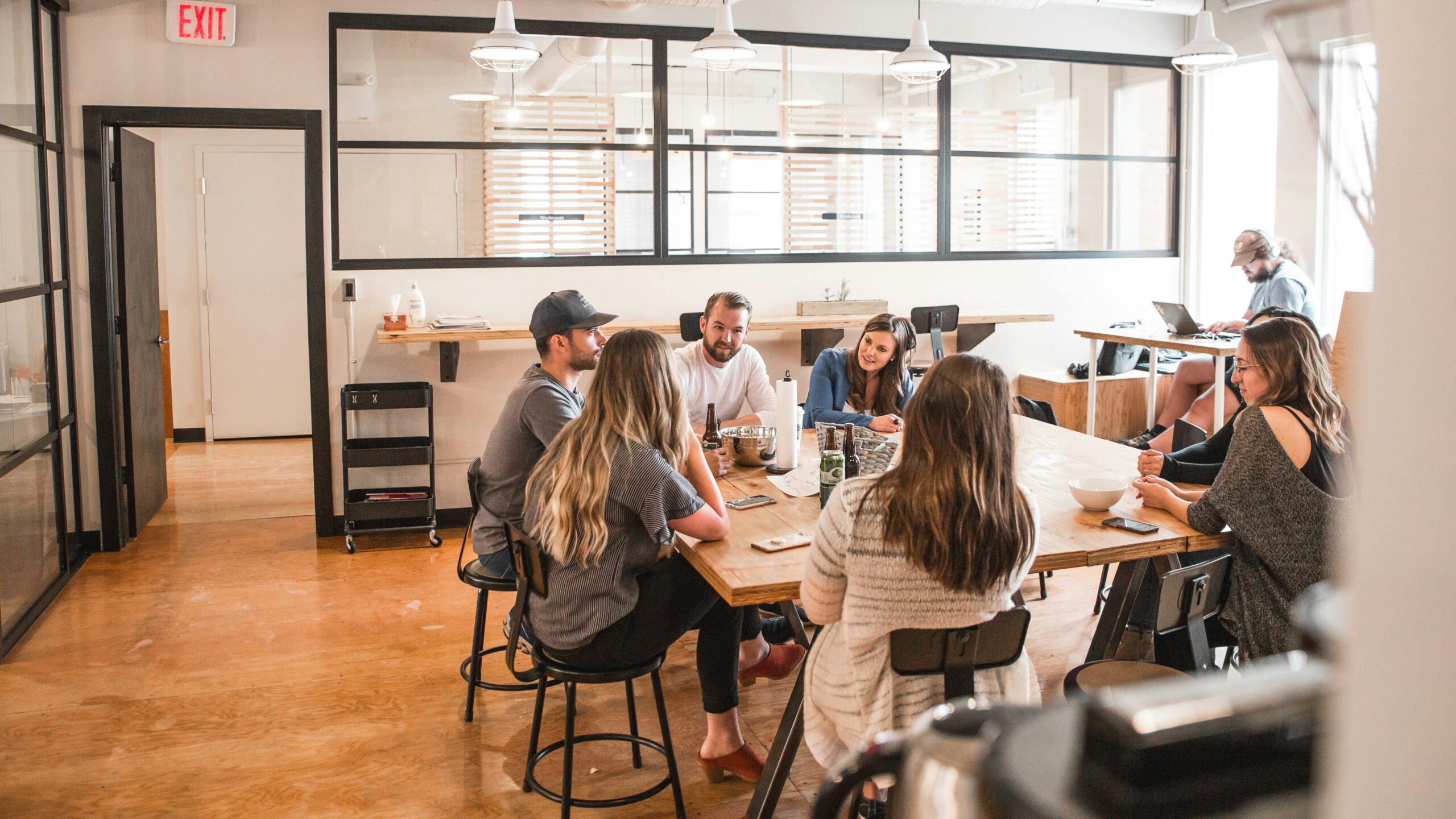 A diverse team of professionals engaged in a meeting around a large wooden table in a bright, modern office with glass partitions.