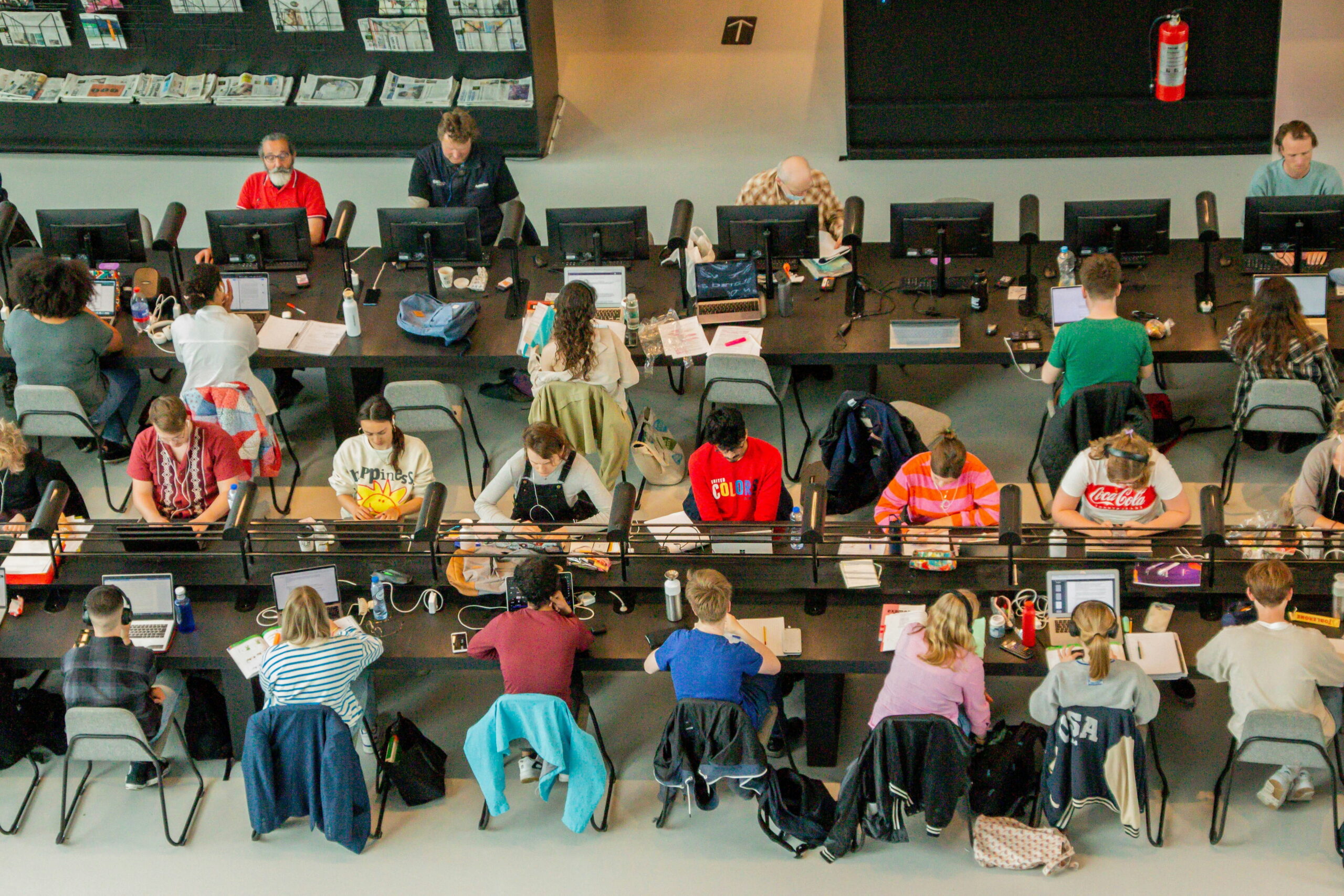 An elevated, high-angle shot of a large, brightly lit co-working room or library filled with multiple rows of desks, computers, laptops, and numerous people working individually or in groups.
