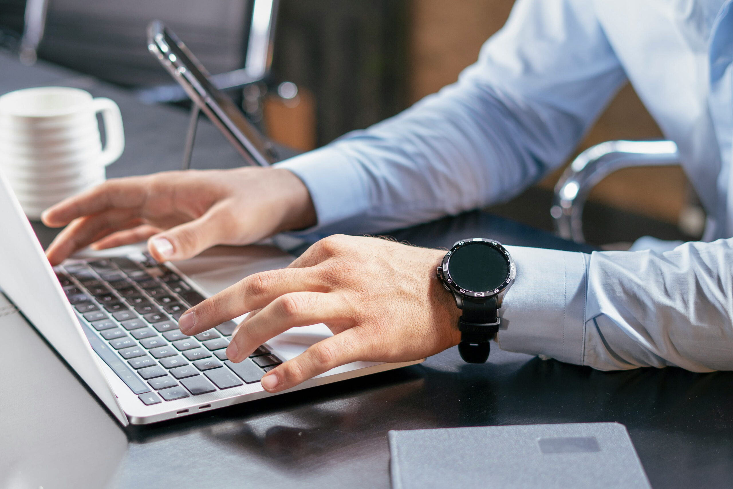 Close-up of a person's hands typing on a laptop keyboard in an office setting, wearing a smartwatch.