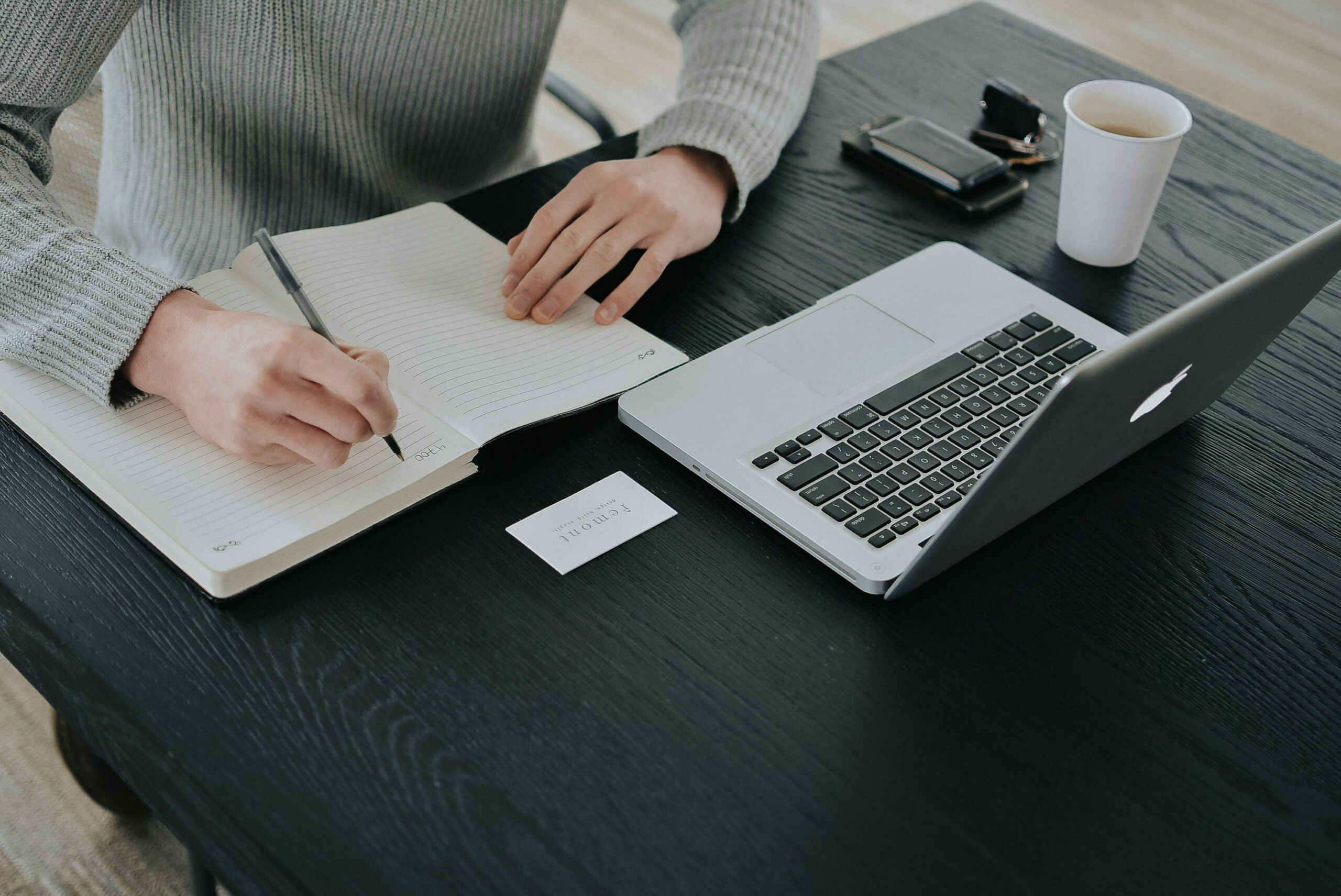 Overhead shot of a person sitting at a black wooden desk, writing in a lined notebook with a pen, next to an open laptop, a coffee cup, and keys.