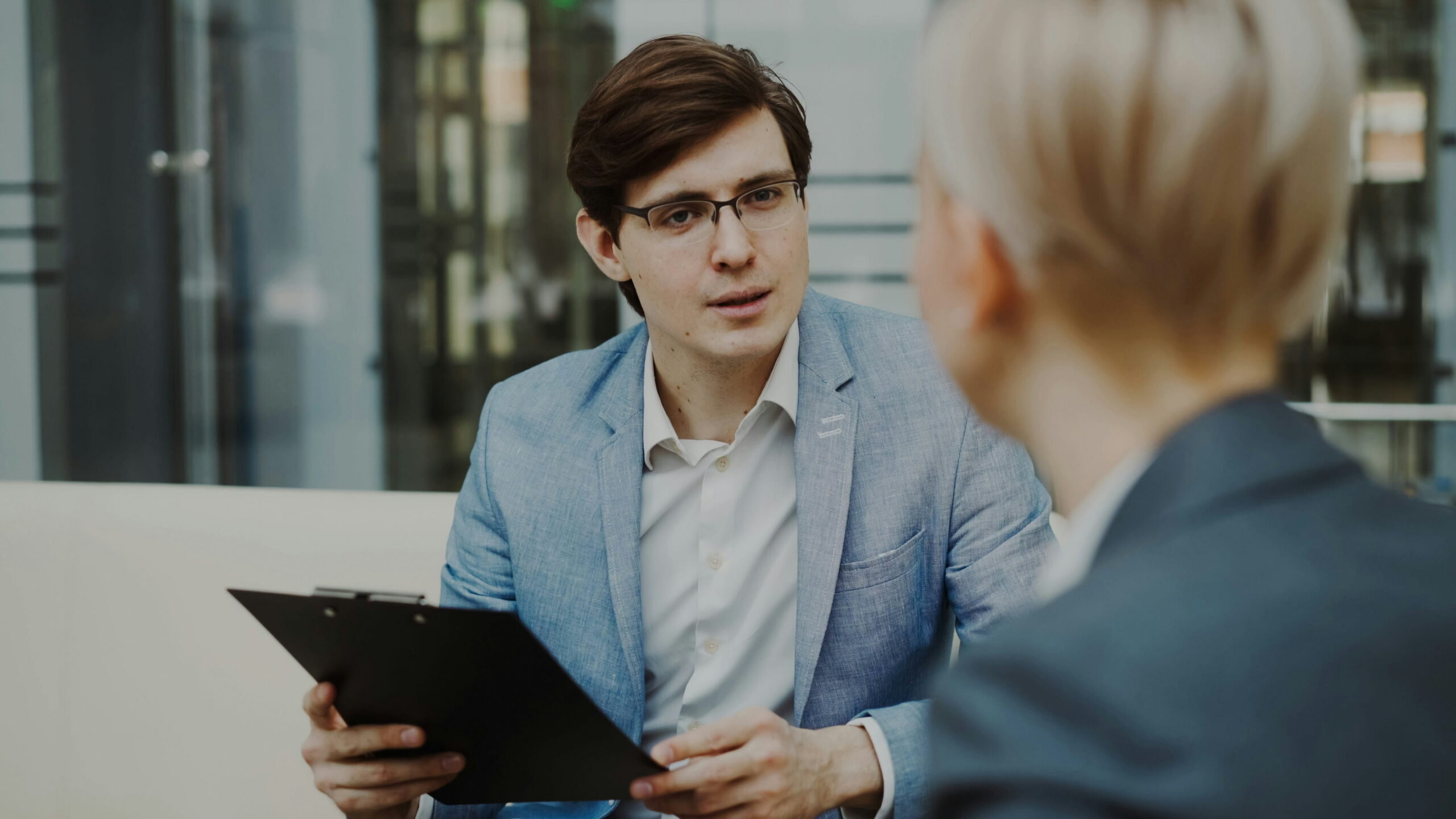 A man in a light blue blazer and glasses holding a clipboard and talking with a colleague during a professional meeting.