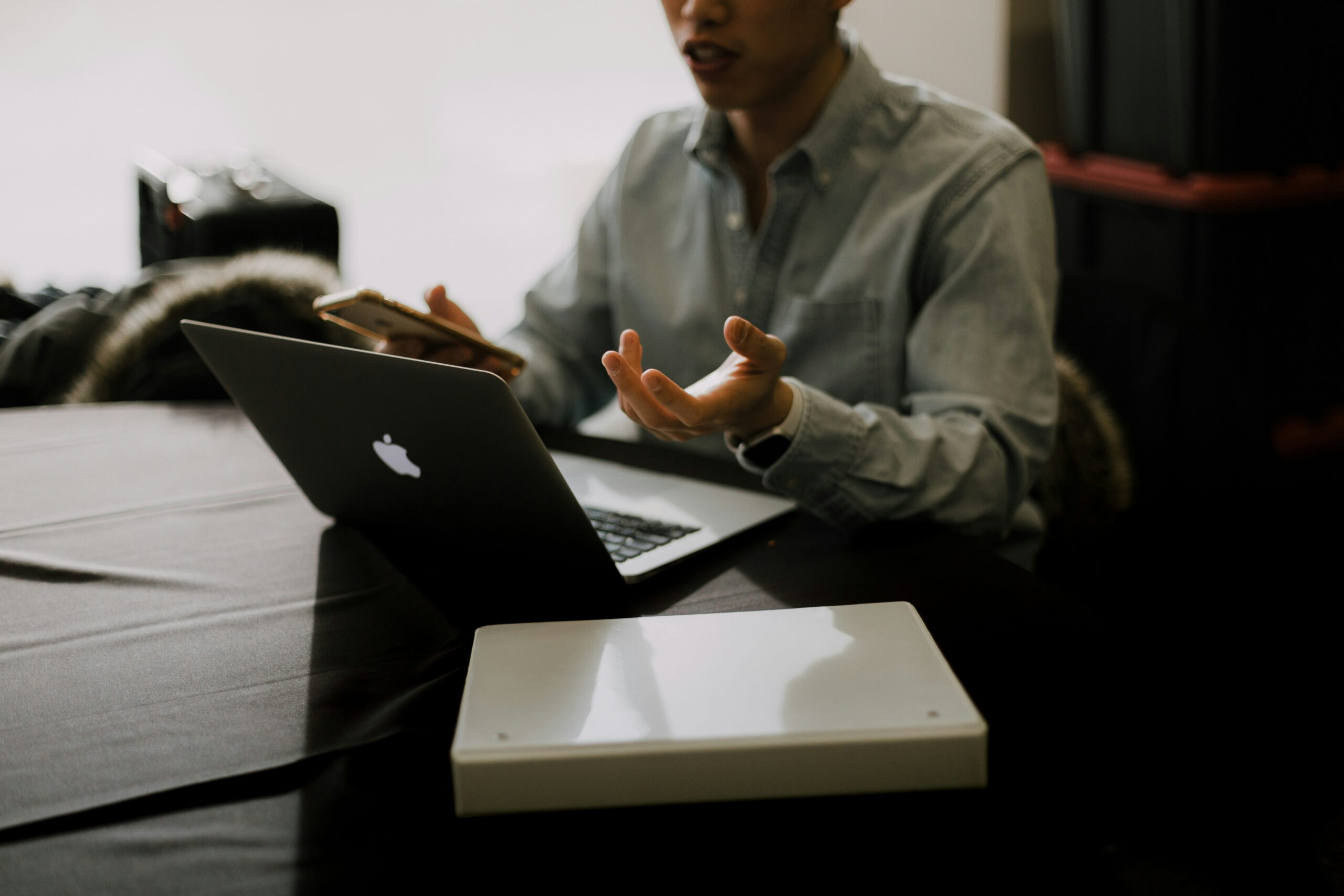 A person sitting at a table with an open laptop and smartphone, gesturing while working.