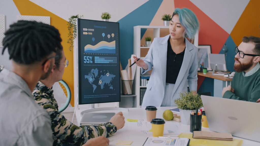 A diverse team of professionals listening as a woman with blue hair points to a computer screen displaying charts, graphs, and a world map in a brightly decorated creative office.