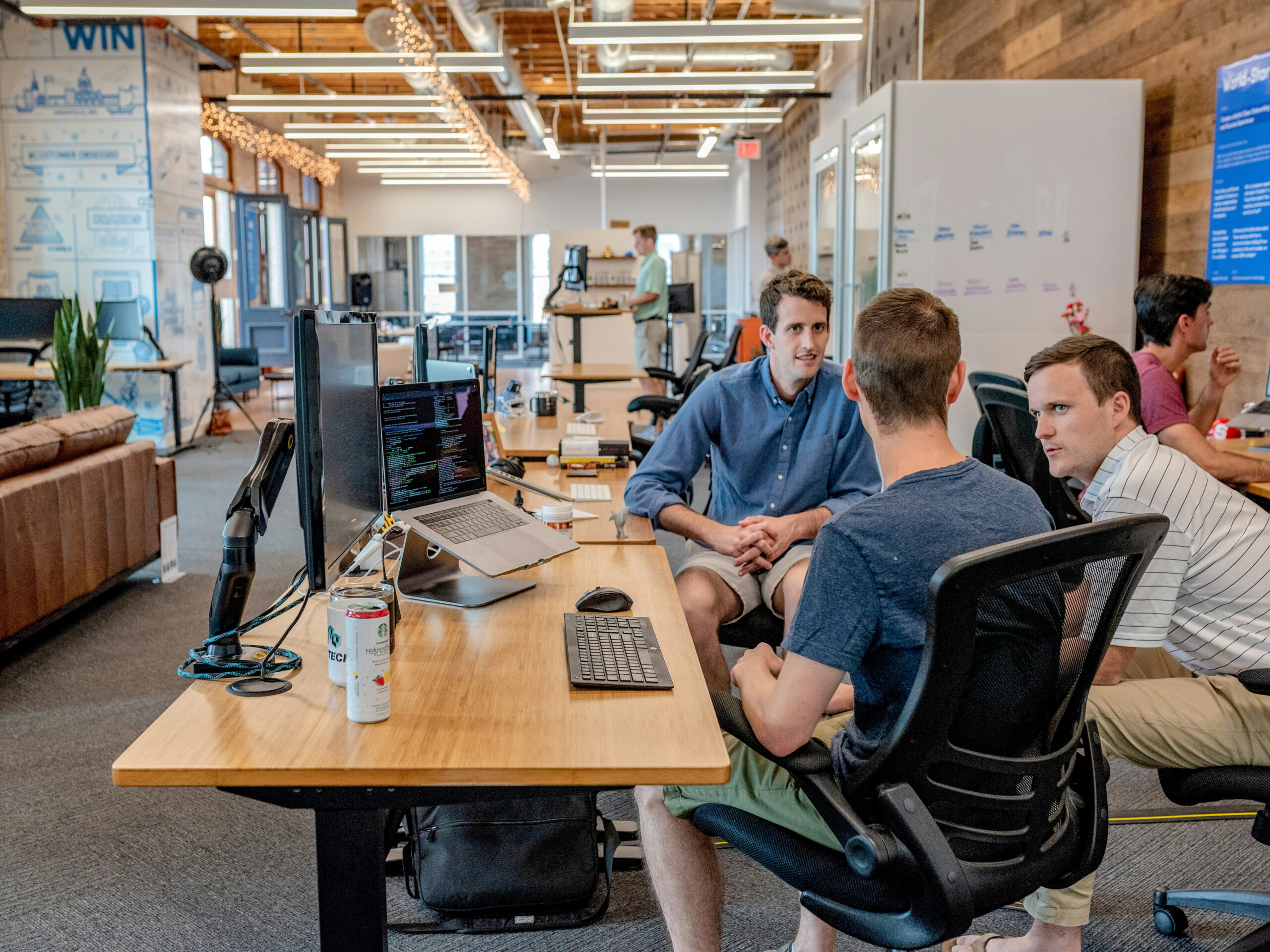 A group of young professionals working and discussing projects together around a long wooden desk in a modern loft office environment.