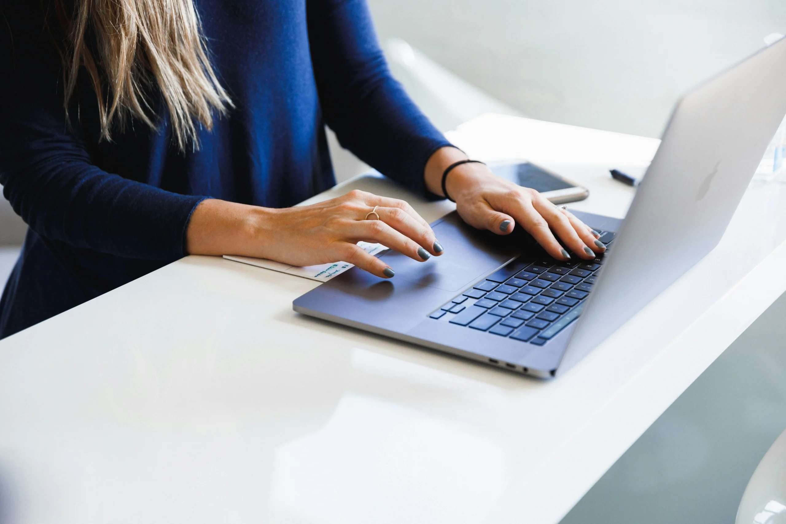 A woman wearing a blue long-sleeve top typing and using the trackpad on a silver laptop resting on a clean white desk.
