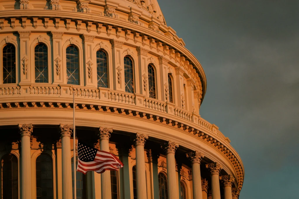 A low-angle shot of the United States Capitol dome and columns with an American flag flying proudly in the foreground at sunset.