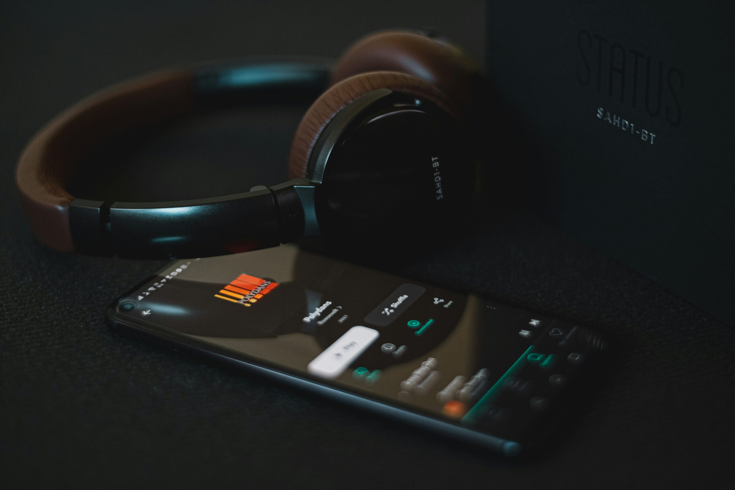 Over-ear wireless headphones lying on a dark surface next to a smartphone displaying an audio player app interface.