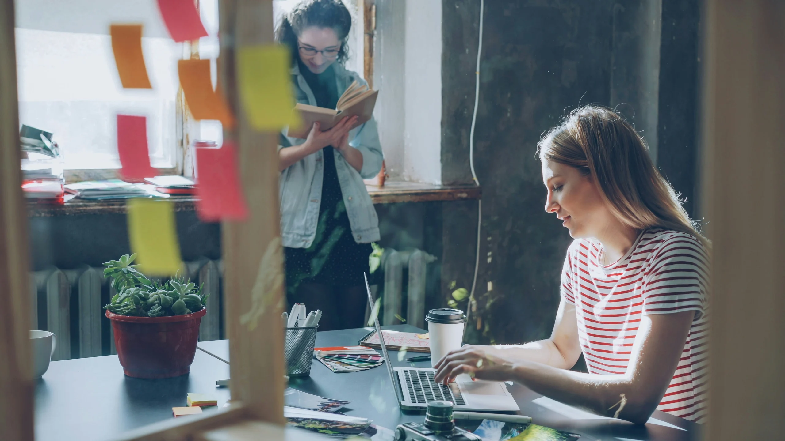 Two young female students working together in a creative study space, with one typing on a laptop at a desk and the other reading a book by the window.