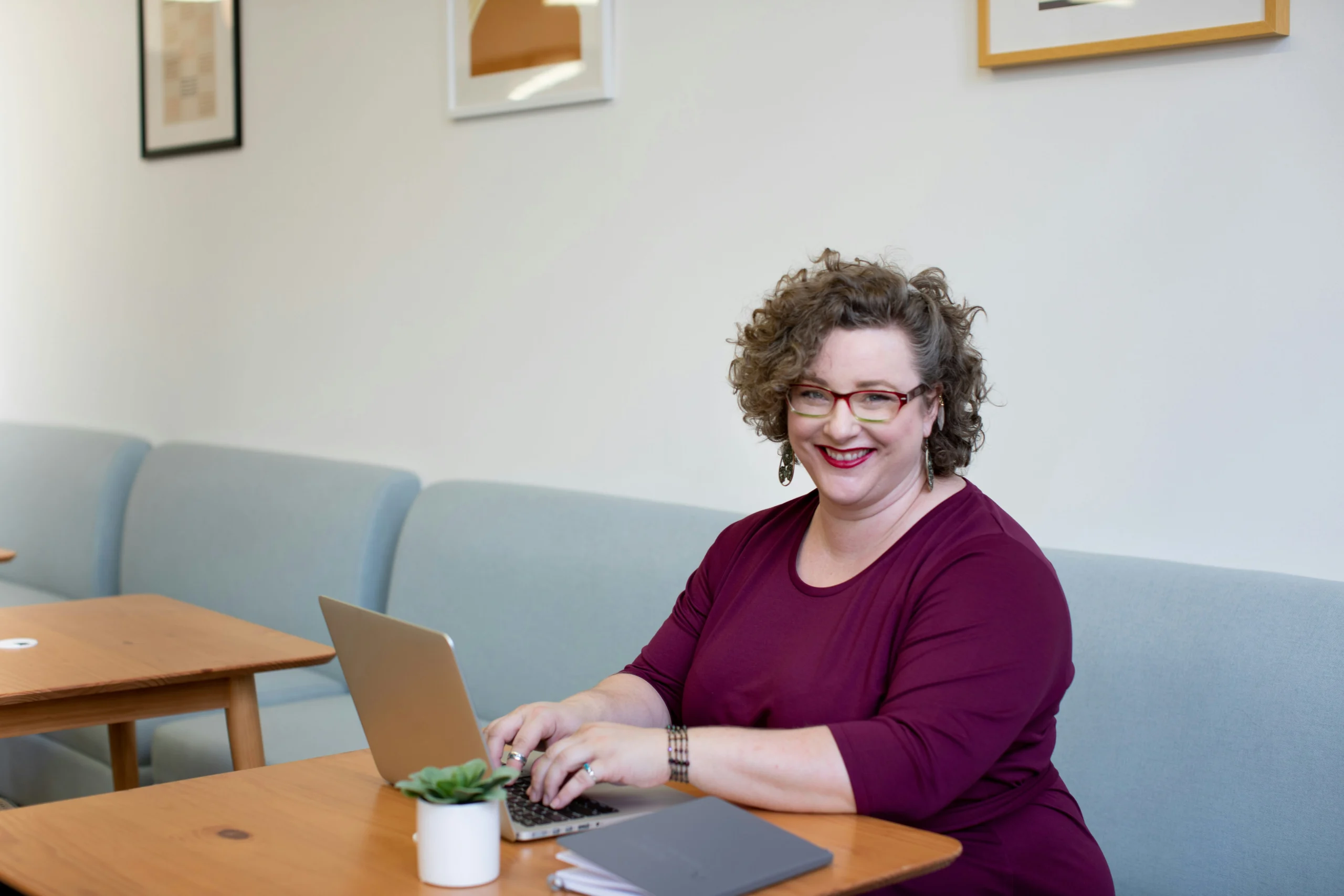 A smiling woman with curly hair and glasses wearing a maroon top, typing on a laptop at a wooden table in a campus or institutional lounge setting.